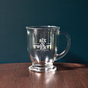 Photo shows glass mug etched with Six & Twenty Distillery logo sitting on wooden table in front of teal wall