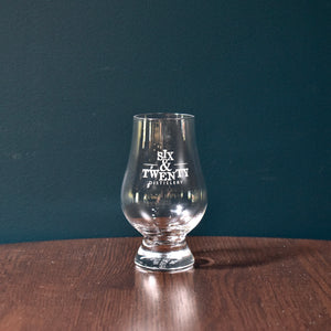 Photo shows a glencairn glass etched with the Six & Twenty logo. The glass has a rounded bowl with slightly flared foot and slightly narrowed neck. Glass sits on a wooden table in front of a teal wall.